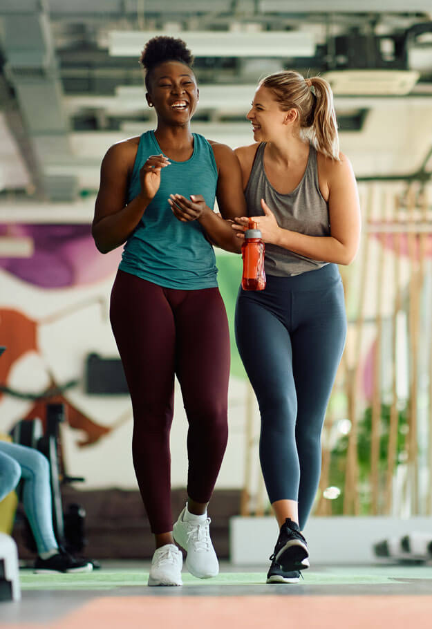 Zwei Frauen gehen lächelnd nebeneinenader her in einem Fitnesstudio und tragen Sportkleidung.